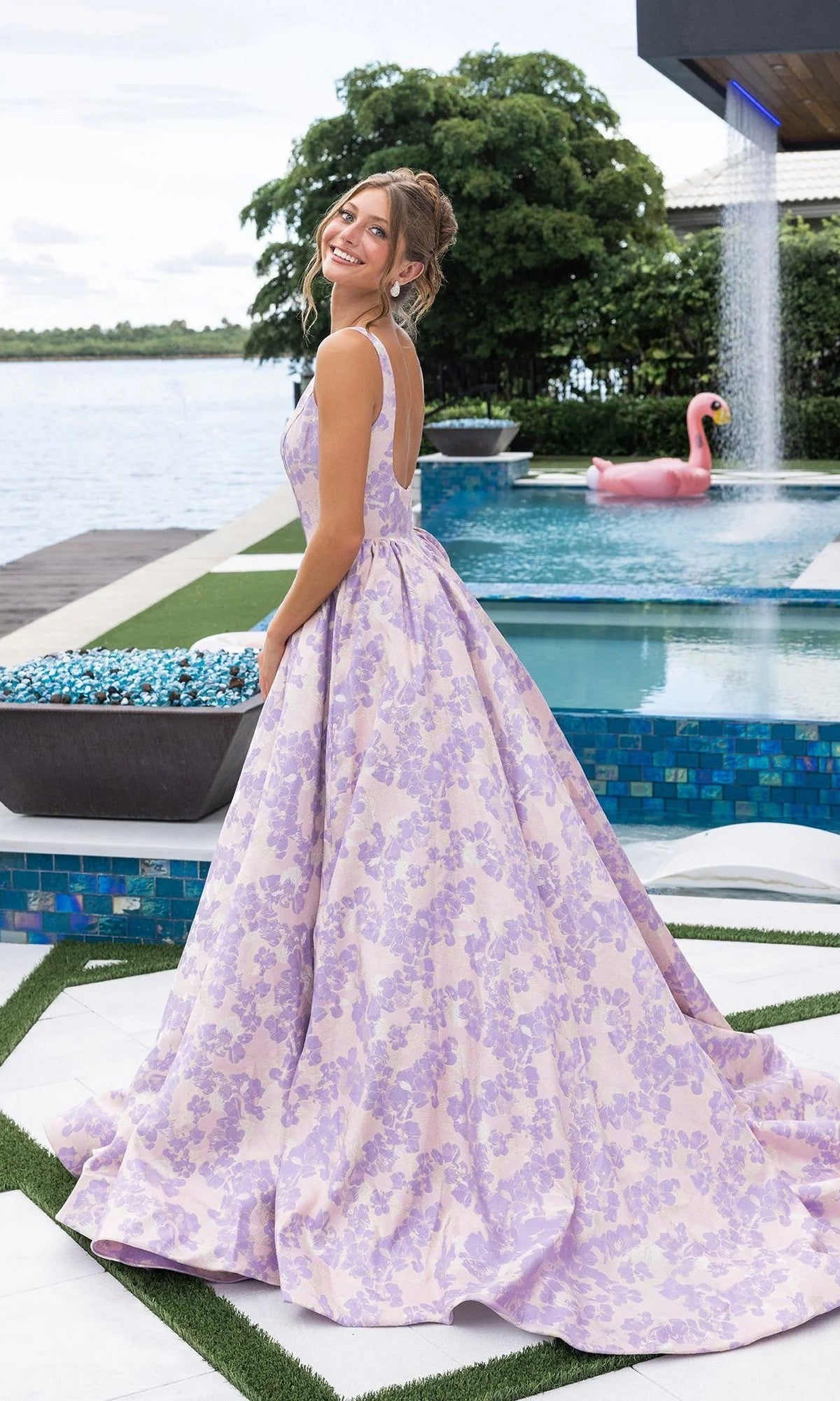 A woman in the Portia and Scarlett XO185 long formal dress smiles over her shoulder by a pool, with a flamingo float and waterfall feature in the background.